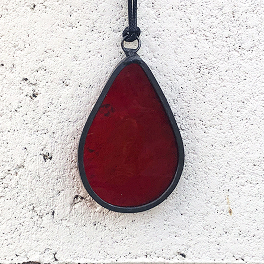 a small red blood drop stained glass charm with translucent red glass against a white wall, no light shining through